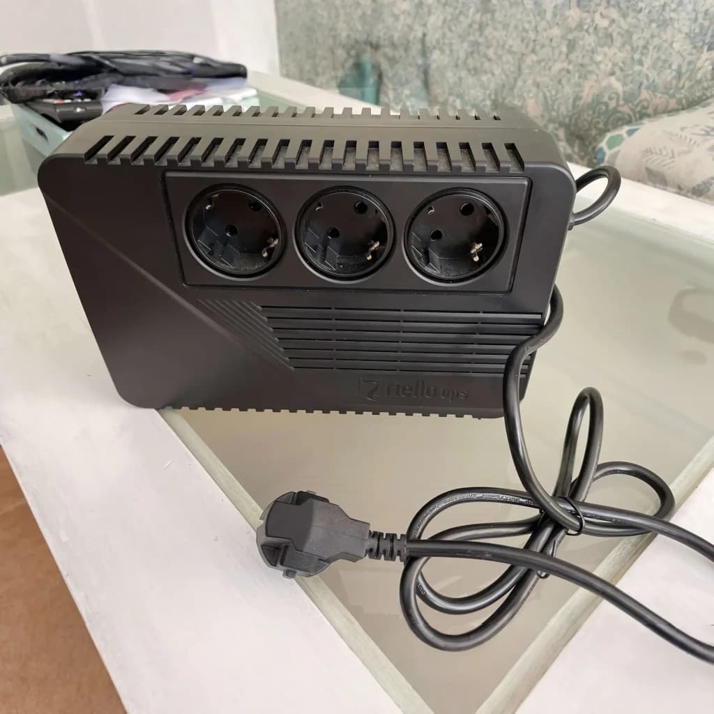 UPS Riello iPlug SE 600VA Linha Interativa 360W Preto, em uso: vista frontal superior sobre mesa, 3 tomadas Schuko e cabo