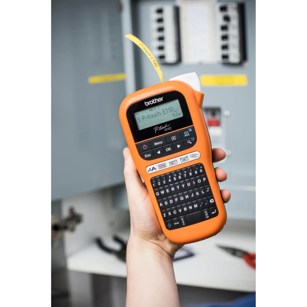 Impresora de etiquetas Brother PT-E110 Negro/Naranja, en uso: mano sostiene el dispositivo frente a cuadro eléctrico, cinta