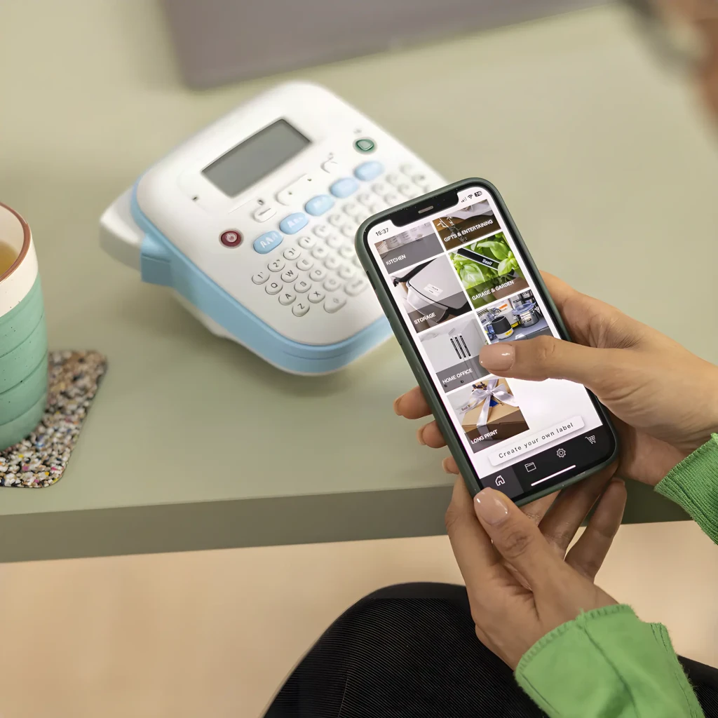 Impresora de etiquetas Brother PT-N25BT Color Bluetooth, en uso: manos con smartphone y app Brother al fondo