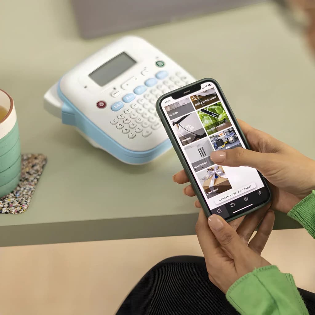 Impresora de etiquetas Brother PT-N25BT Color Bluetooth, en uso: manos con smartphone y app Brother al fondo