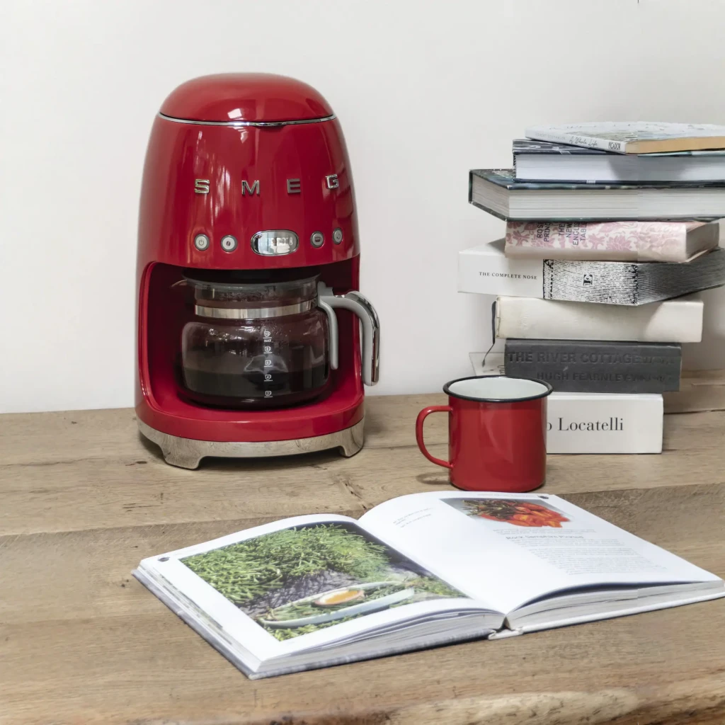 Cafetera de goteo Smeg 1,4L 1050W Rojo DCF02RDEU, en uso: sobre mesa de madera con jarra llena, taza roja y libros apilados