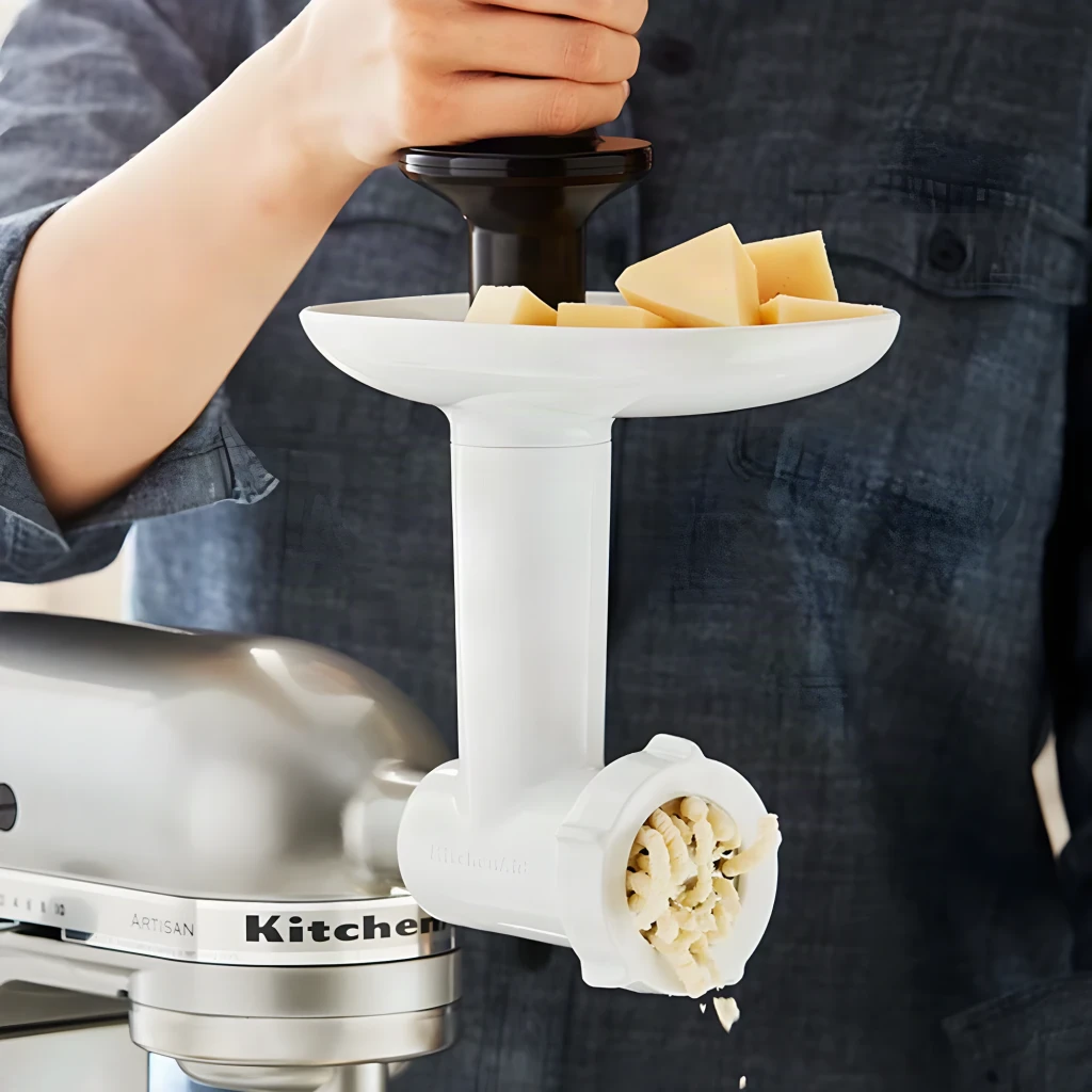 Picador de carne KitchenAid Blanco 5KSMFGA, en uso: manos introduciendo queso en la bandeja, queso rallado saliendo