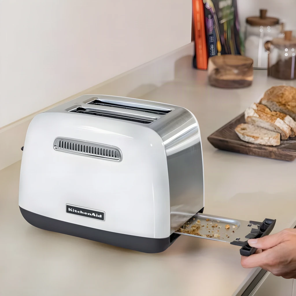 Tostadora KitchenAid 1100W 2 Ranuras Blanco 5KMT2115EWH, en uso: mano extrayendo bandeja de migas en encimera con pan