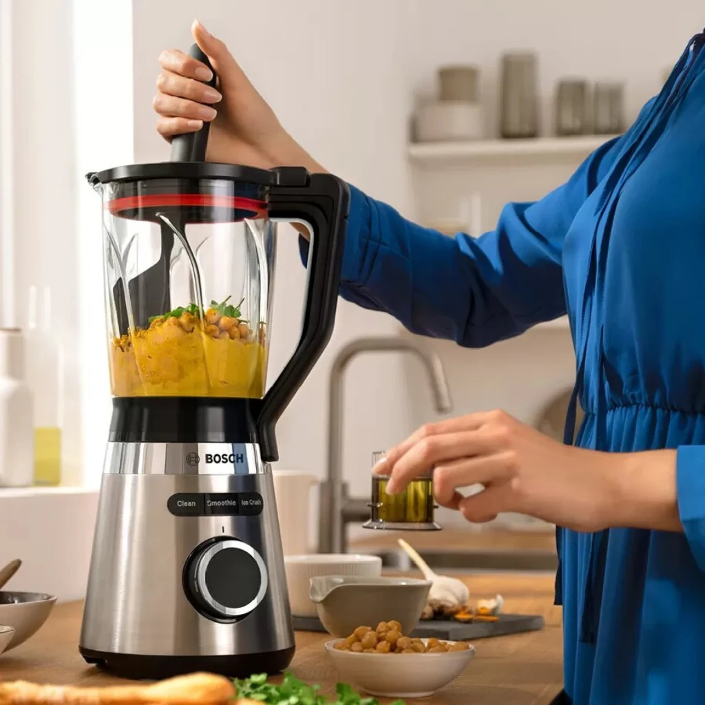 Batidora de vaso Bosch VitaPower 1.5L 1200W Negro MMB6382M, en uso: mujer presiona la tapa mientras tritura hummus en cocina