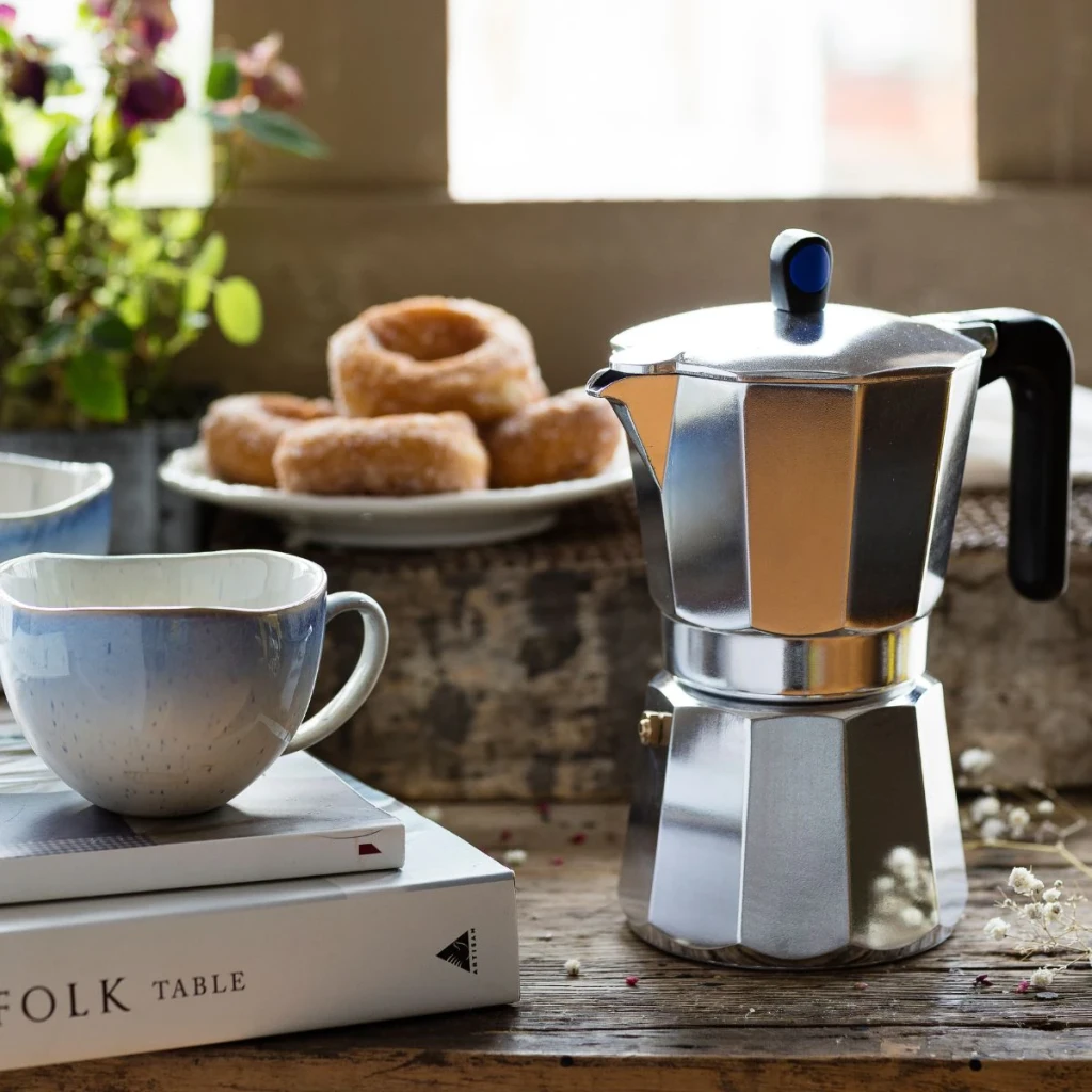 Cafetera italiana Monix Induction Express 6 Tazas Plata M860006, en uso: sobre mesa rústica con taza y donuts al fondo