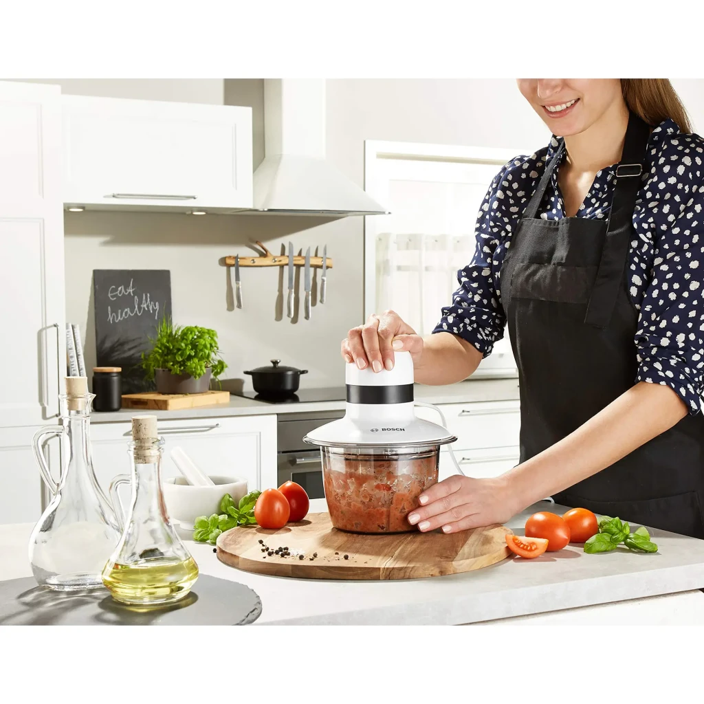 Hachoir Bosch 0,8L 400W Blanc MMR08A1, en utilisation: femme hachant des tomates sur planche en bois en cuisine moderne