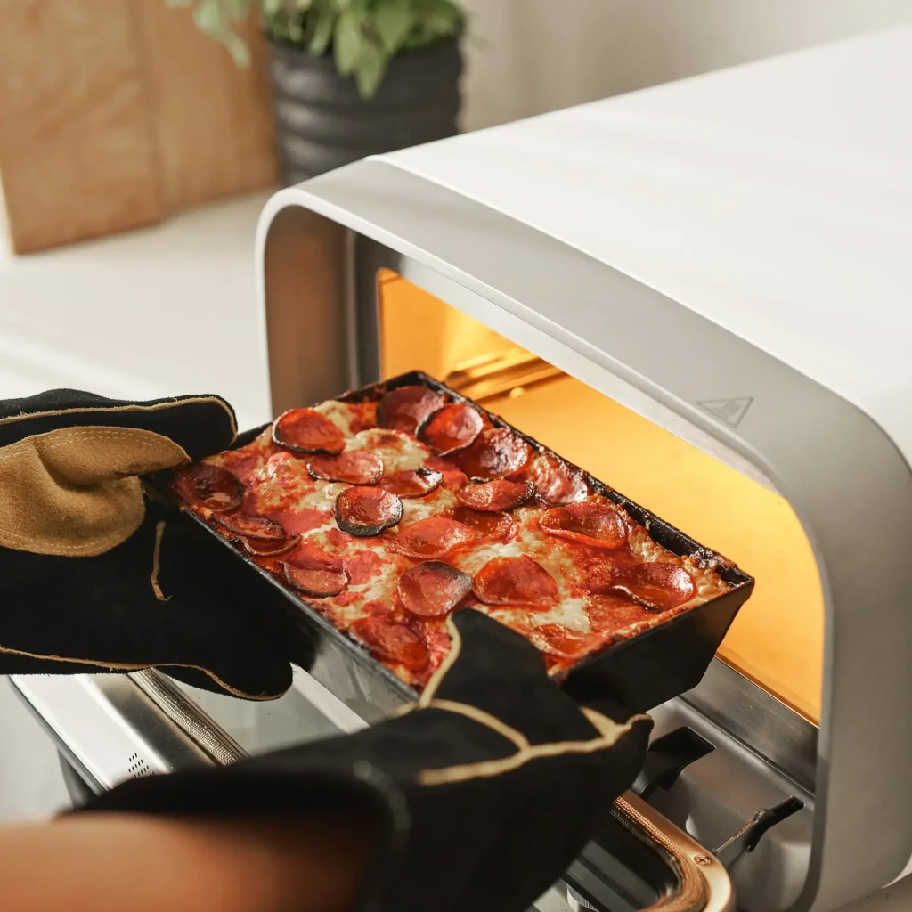 Four à pizza électrique Ooni Volt 2 1600W Blanc, en utilisation: mains gantées sortent une pizza pepperoni dans un moule