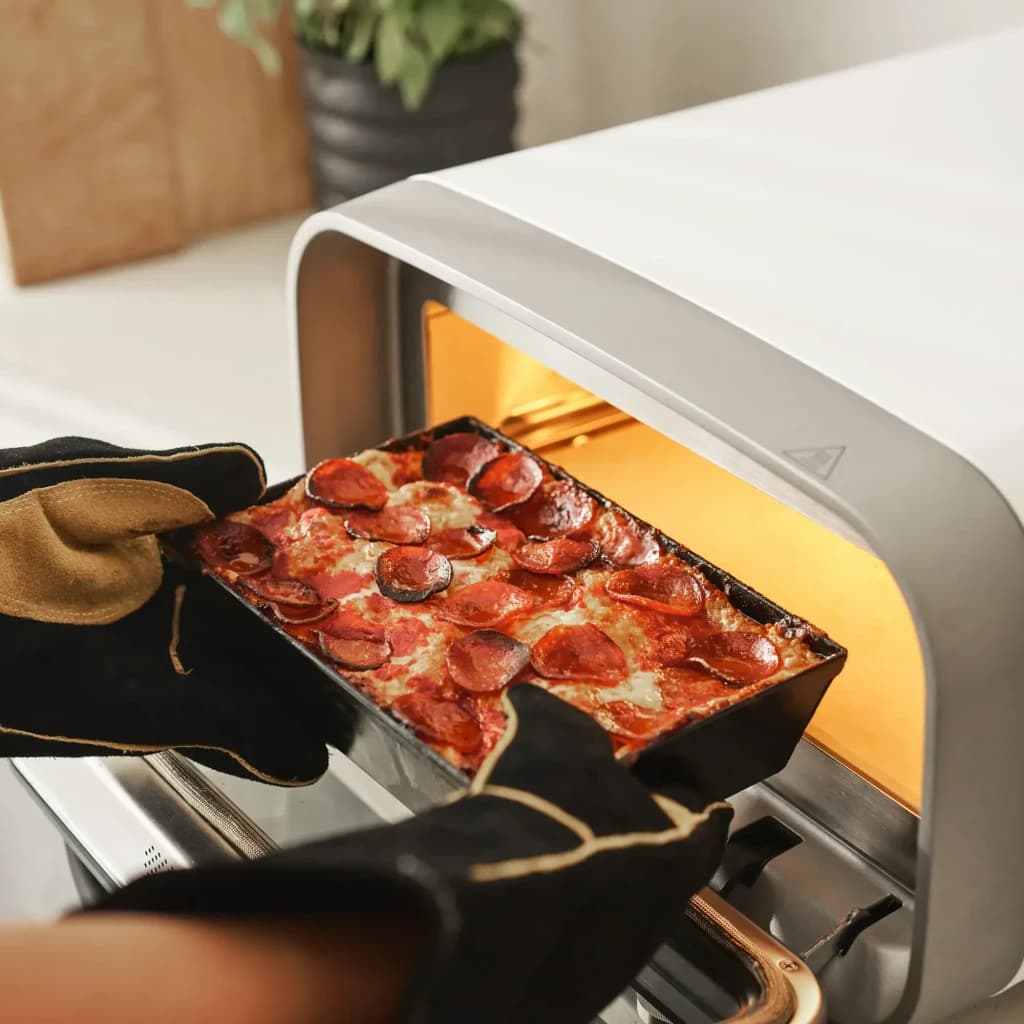 Four à pizza électrique Ooni Volt 2 1600W Blanc, en utilisation: mains gantées sortent une pizza pepperoni dans un moule