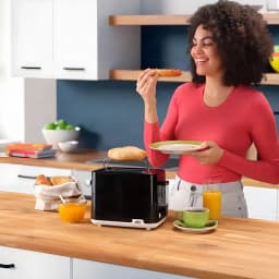 Grille-pain Braun HT 1010 900W 2 Fentes Noir, en utilisation: femme souriante au petit-déjeuner avec toasts et jus