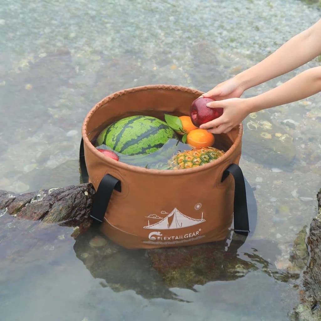 Seau pliable Flextail Flex Bucket 20L Marron 32x25x32cm, en utilisation: fruits rafraîchis en rivière, mains saisissant pomme
