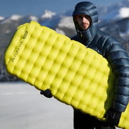 Colchão Insuflável Flextail Tiny R09 AVS Múmia L Verde Valor R 9,4, em uso: homem com casaco de penas segura o colchão