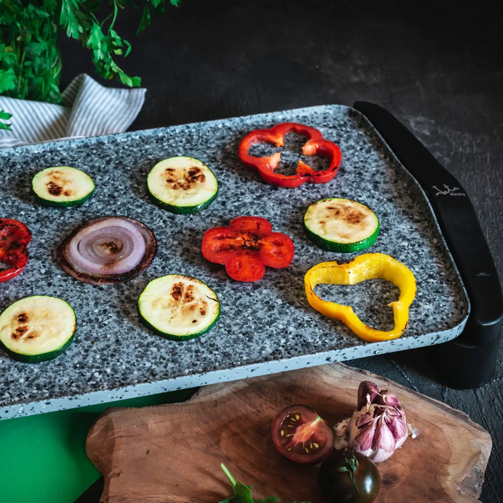 Plancha de asar JATA GR558 2500W Negro/Gris, en uso: superficie de piedra con verduras a la plancha, pimientos, calabacín