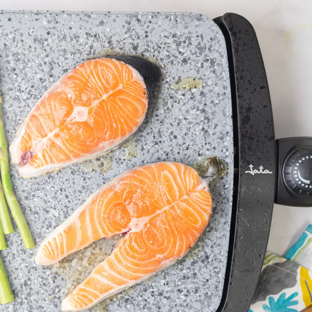 Plancha de asar JATA GR558 2500W Negro/Gris, en uso: dos rodajas de salmón y espárragos sobre superficie efecto piedra