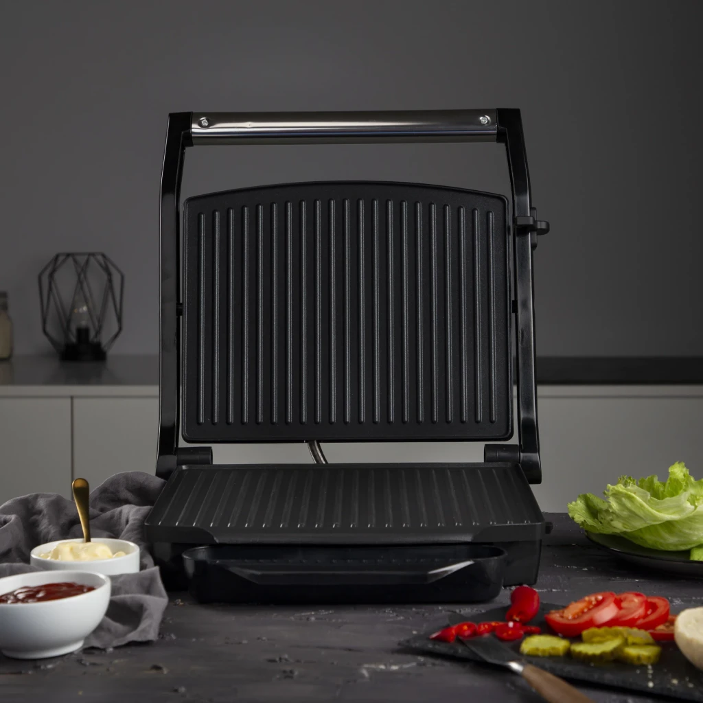 Princess Panini Grill 2000W Plata/Negro 112415, en uso: tapa abierta con placas estriadas, rodeado de tomates, pepinillos