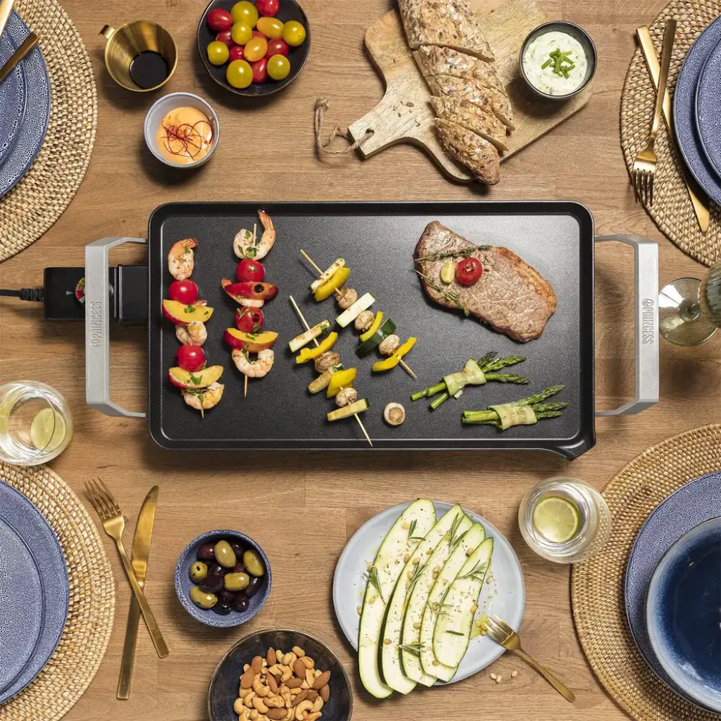 Plancha de asar Princess Table Chef Premium XL 2500W 460x260mm Negro, en uso: vista cenital sobre mesa con brochetas
