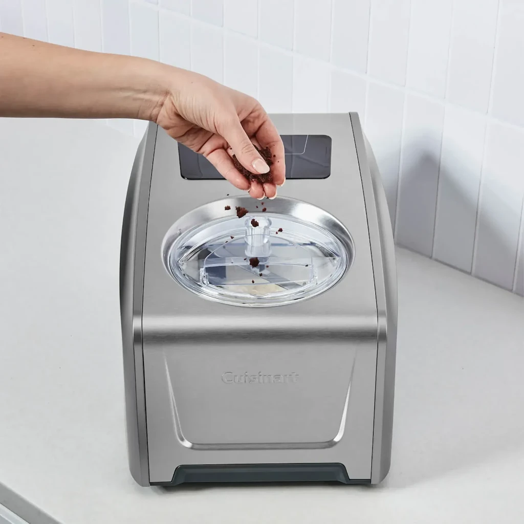 Sorbetière Cuisinart 1,5L Acier Inoxydable ICE150E, en cours d'utilisation: main ajoutant des ingrédients par le couvercle