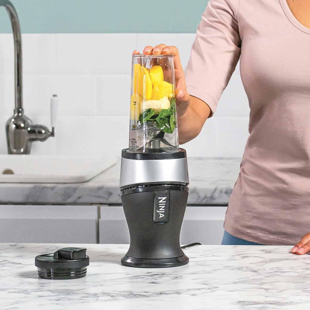 Liquidificador Ninja 0,47L 700W Preto/Prata QB3001, uso em cozinha: mulher coloca copo com frutas e espinafre na base