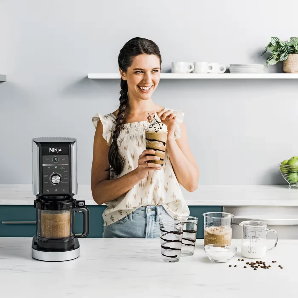 Heladora Ninja CREAMi Deluxe 0,709L 800W Negro NC502EU, en uso: mujer sostiene milkshake con nata junto a la máquina