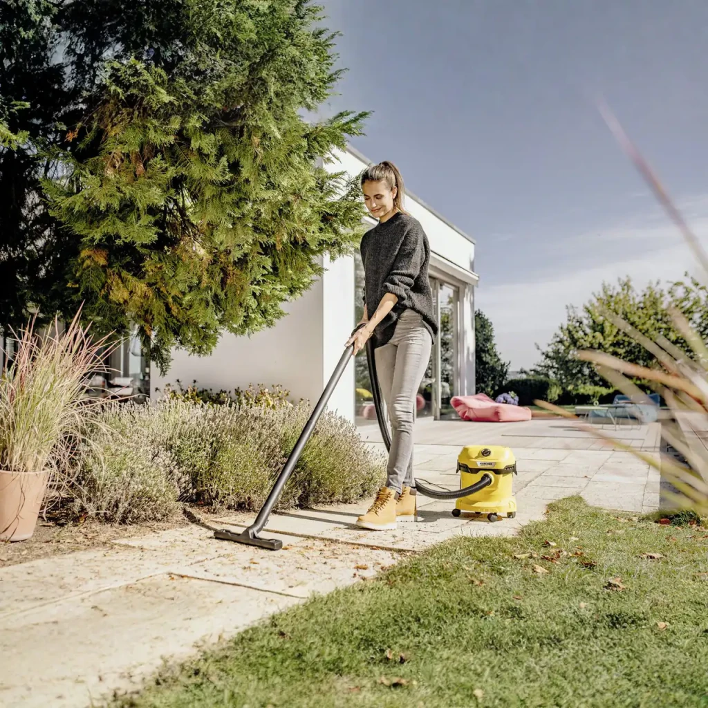 Aspirador en seco y húmedo Kärcher WD 2 Plus Amarillo 1000W V-12/4/18, en uso: mujer aspirando terraza exterior con jardín