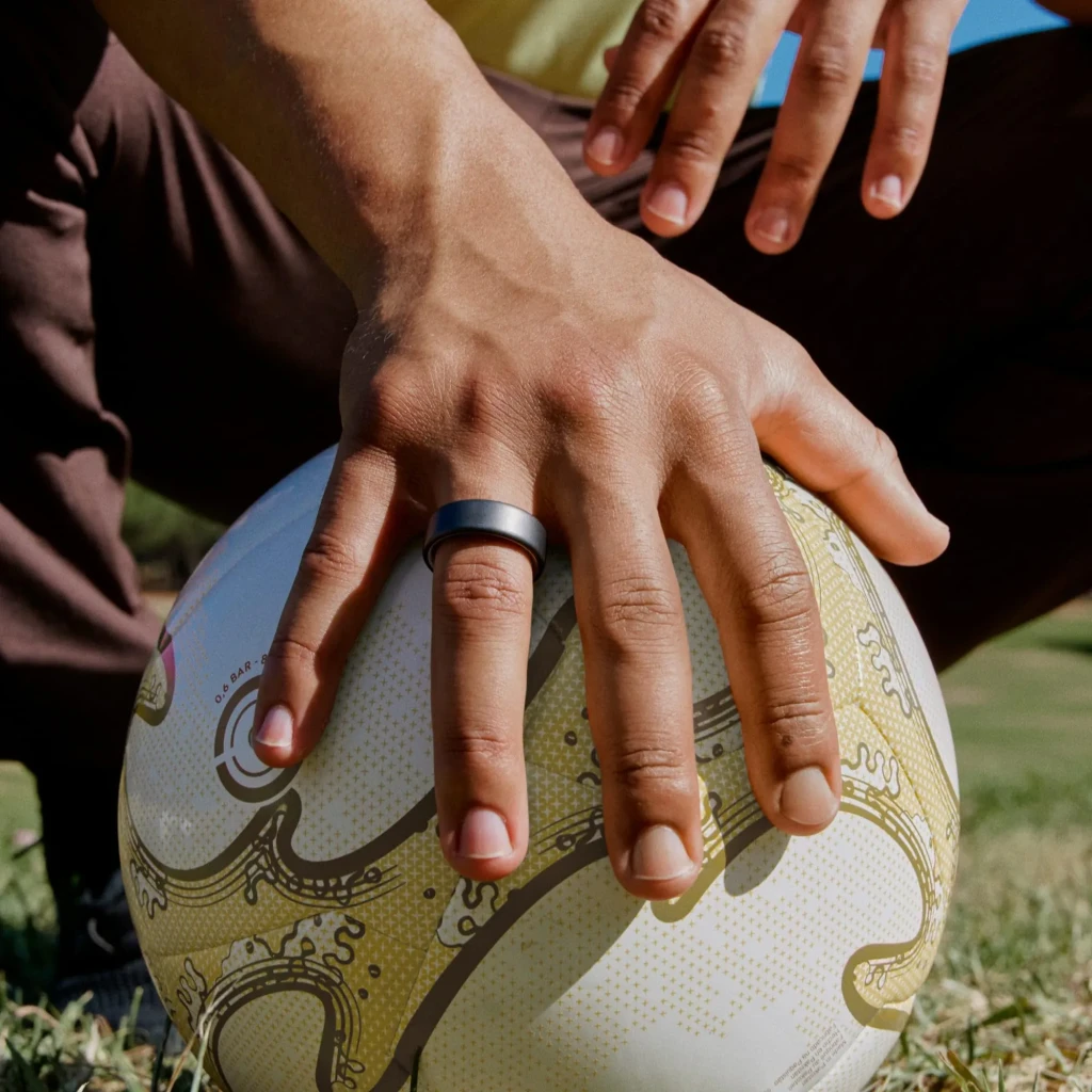 Oura Ring 4 – Anel Inteligente, Tamanho 8, Preto, em uso: anel preto no dedo indicador segurando bola de râguebi ao ar livre