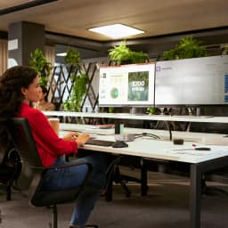 Combo Teclado y Ratón HP 725 Negro Inalámbrico Español, en uso: mujer tecleando en oficina con dos monitores y plantas