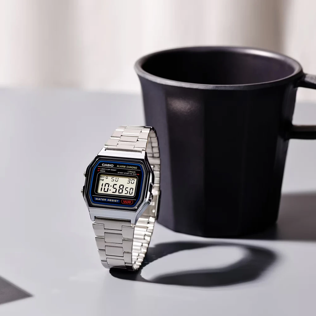 Casio A158WA-1DF Vintage Digital Cronómetro, Alarme, Calendário, em uso: relógio prateado apoiado em mesa junto a caneca