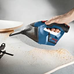 Aspirador de mão Bosch GAS 12V Preto/Azul Sem Fios 900l/min, em uso: mão aspirando serradura de madeira em superfície
