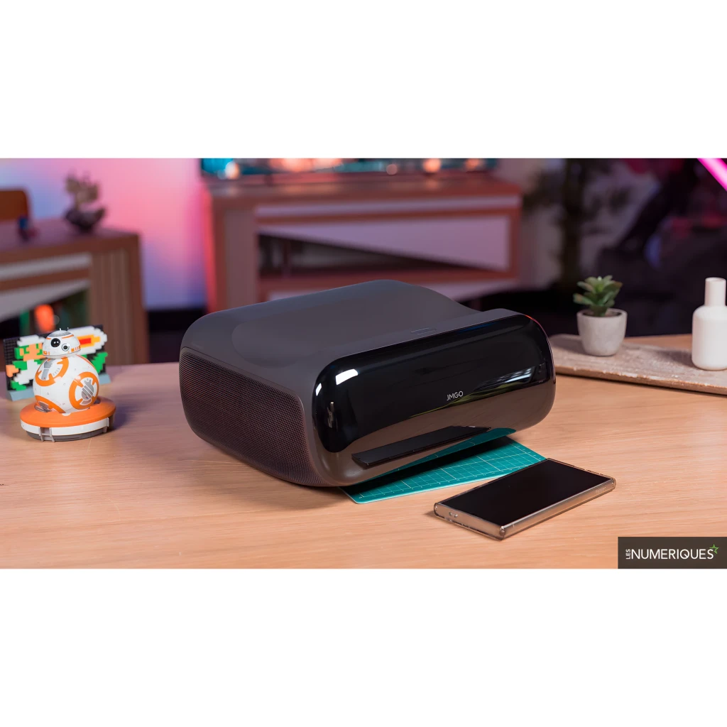 JMGO Proyector O2S Ultra 64GB negro, vista lateral en salón: sobre mesa de madera junto a smartphone y figura BB-8