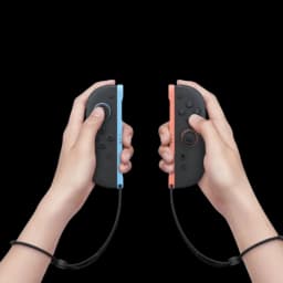Nintendo Switch 2 Joy Con en manos, uno gris con borde azul y otro negro con borde rojo, botones visibles y correas de muñeca conectadas