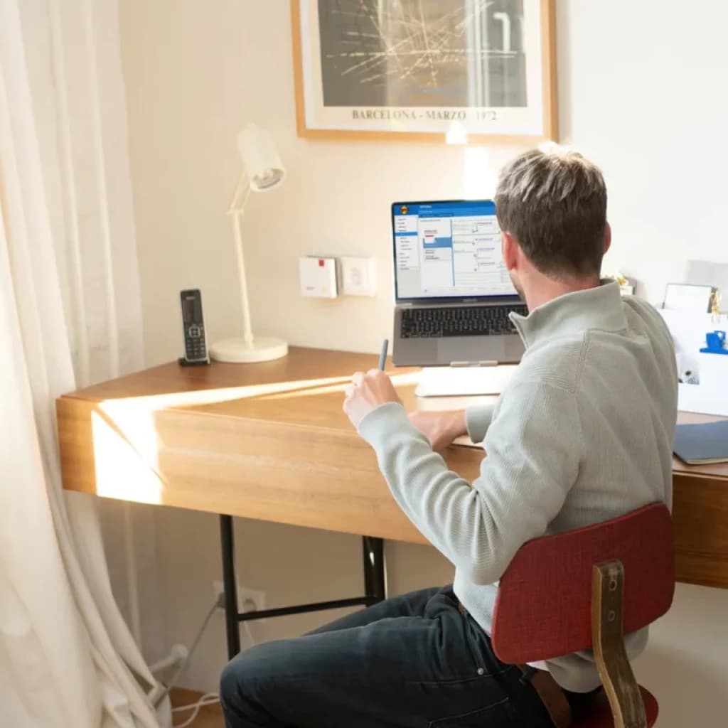 Repetidor FRITZ! 1200 AX branco na parede, LEDs de status visíveis ao lado de uma mesa de madeira com laptop, lâmpada e telefone, ambiente doméstico moderno