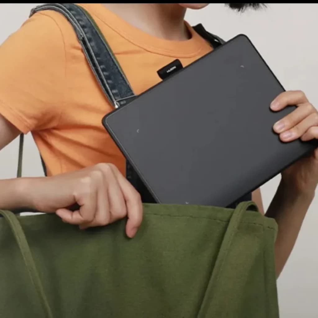 Tablette graphique compacte gris anthracite rangée dans un sac vert, bords arrondis, surface lisse, format portable; main tenant la tablette par le côté