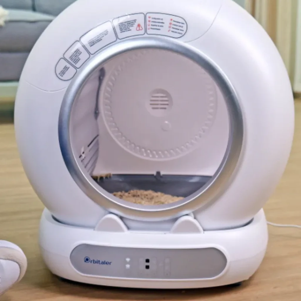 Vista frontal do Orbitaler Yupiter AstroClean 6700 Smart Litter Ultra Compact com gato visto de frente e smartphone com aplicação compatível - Item1