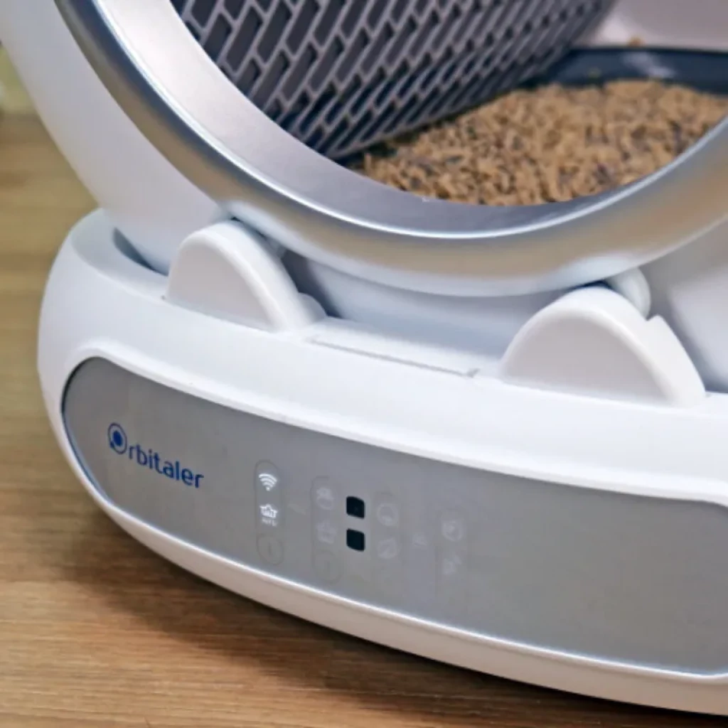 Vista frontal do Orbitaler Yupiter AstroClean 6700 Smart Litter Ultra Compact com gato visto de frente e smartphone com aplicação compatível - Item2