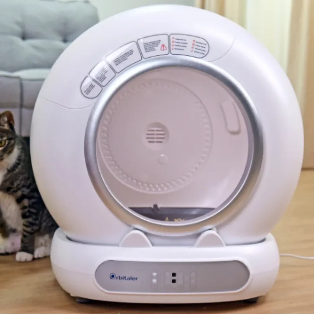 Vista frontal do Orbitaler Yupiter AstroClean 6700 Smart Litter Ultra Compact com gato visto de frente e smartphone com aplicação compatível - Item4